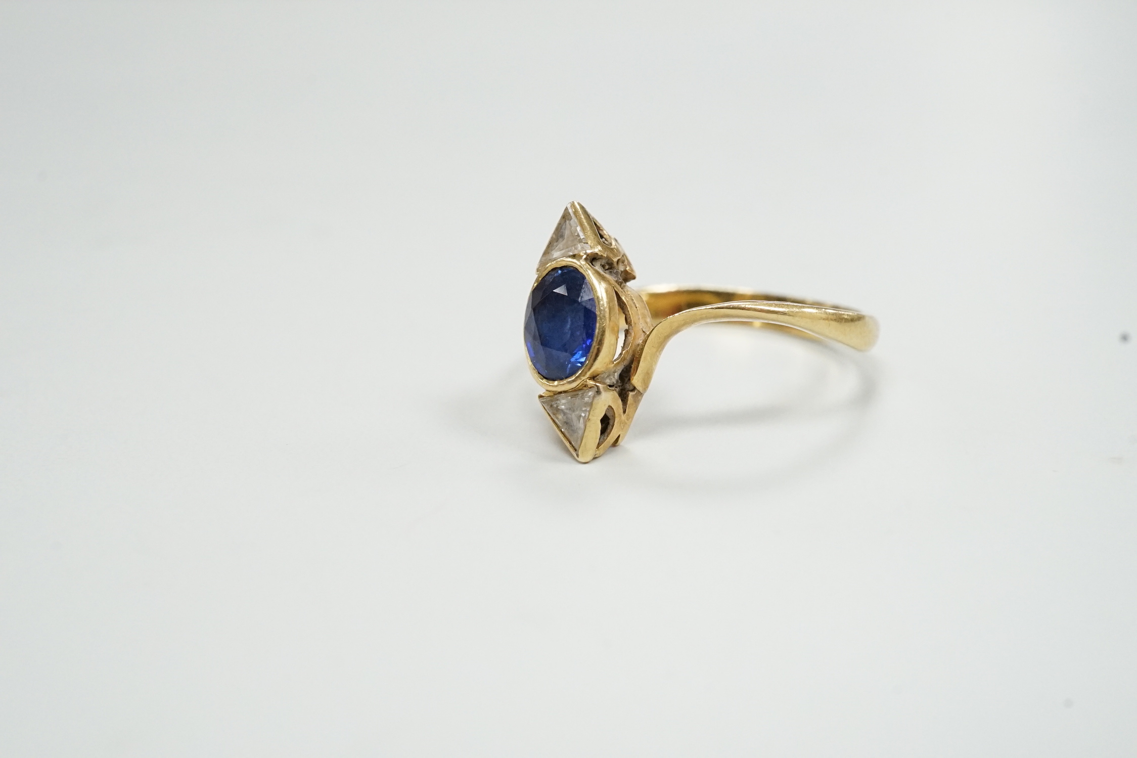 A yellow metal and single stone oval cut sapphire and two stone triangular cut diamond set ring, size L/M, gross weight 2.4 grams.